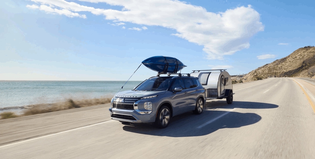 Mitsubishi Outlander PHEV 2025 gris remorquant une mini-roulotte sur une route côtière, avec un kayak sur le toit, illustrant ses capacités de remorquage et d'aventure en mode hybride rechargeable.